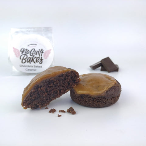 Two chocolate baked goods topped with caramel, one of which is cut in half, displayed next to a package labeled "No Guilt Bakes Chocolate Salted Caramel" and a few pieces of dark chocolate.