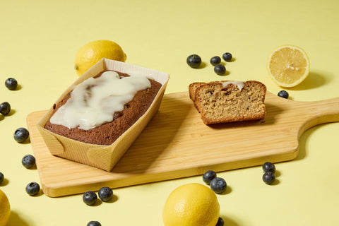 A loaf cake in a cardboard tray with white icing sits on a wooden board alongside a sliced piece. Surrounding them are whole and halved lemons, and scattered blueberries, all on a yellow background.