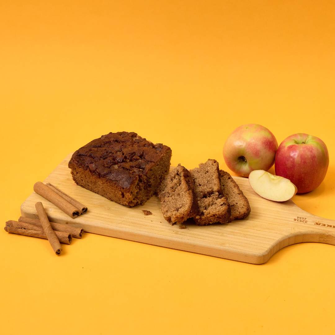A wooden cutting board displaying a loaf of sliced bread, whole apples, apple slices, and cinnamon sticks against a solid orange background evokes the essence of preparing No Guilt Bakes' Apple and Cinnamon Keto Cake Loaf | No Added Sugar | Low Carb.