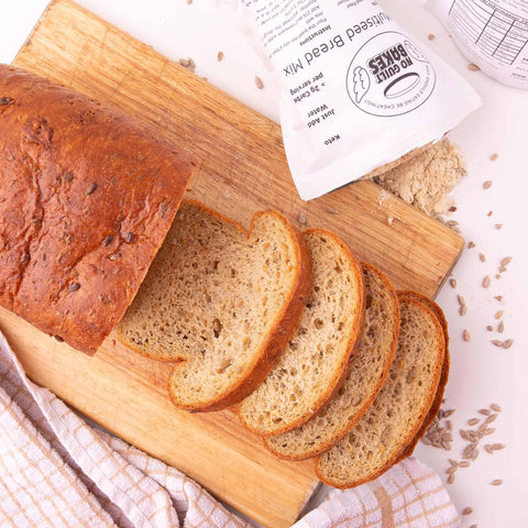 Sliced whole grain bread from The Regular Bread Bundle on a wooden cutting board with a pancake mix package and grains nearby. Brand Name: No Guilt Bakes