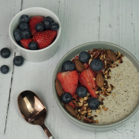 A bowl of chia pudding topped with strawberries, blueberries, almonds, and granola, accompanied by a spoon and a small bowl of mixed berries on a wooden surface. Ideal for a low-carb diet. Made with No Guilt Bakes' Regular Bread Bundle.