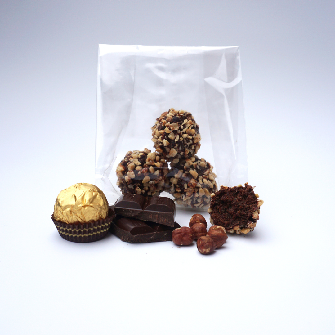 A selection of chocolates, featuring a individually wrapped candy, a chocolate piece with nuts, and hazelnuts, are displayed in front of a transparent bag containing three Hazelnut and Chocolate Snowballs from No Guilt Bakes.