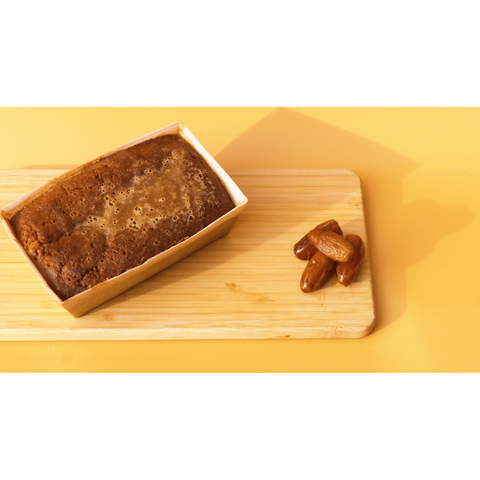 Placed on a wooden board, a Sticky Toffee Keto Cake Loaf by No Guilt Bakes rests in a paper tray beside a small pile of dates, all on a vibrant yellow background. It's an irresistible keto-friendly delight for anyone searching for a low-carb dessert option.