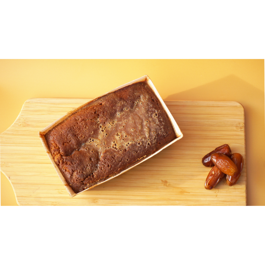 A loaf of Sticky Toffee Keto Cake from No Guilt Bakes sits in a rectangular wooden tray on a wooden board, accompanied by a few whole dates, providing a delightful low-carb dessert option.