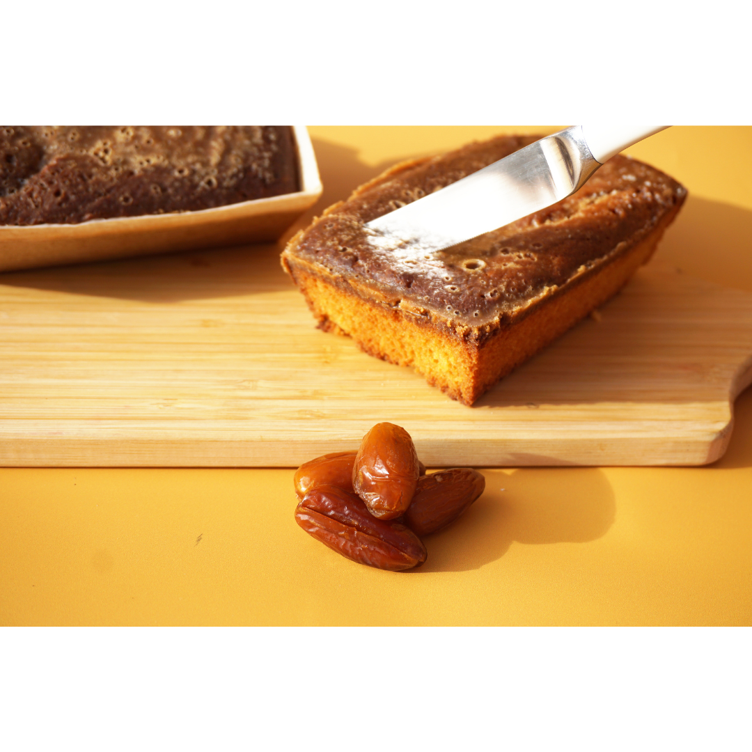 A Sticky Toffee Keto Cake Loaf from No Guilt Bakes sits on a wooden cutting board with a knife resting on top, accompanied by a few dates; this keto-friendly treat makes for a delightful low-carb dessert.