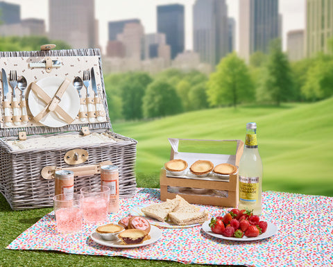 A picnic setup with a wicker basket, sandwiches, strawberries, No Guilt Bakes Strawberry Tarts (Low Carb and Keto), soda cans, and lemonade on a colorful blanket. City buildings and green trees are visible in the background.