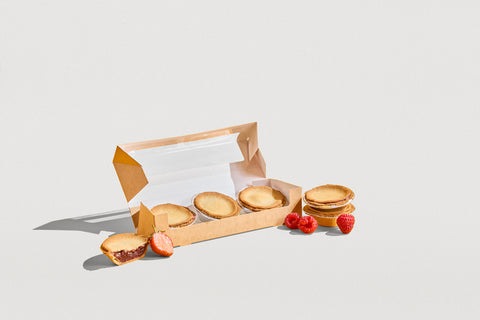 A partially open brown paper box from No Guilt Bakes contains several gluten-free tartlets, including low-carb dessert options and a few raspberries. A Strawberry Tart from their keto-friendly 6 Tarts collection is placed alongside it on a white surface.