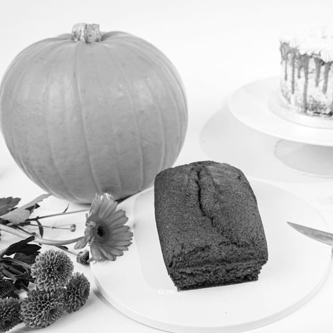 A gluten-free pumpkin, a loaf of bread, a Pumpkin and Caramel Keto Cake Loaf by No Guilt Bakes | No Added Sugar | Low Carb | Limited Edition, and some flowers are arranged on a white surface.