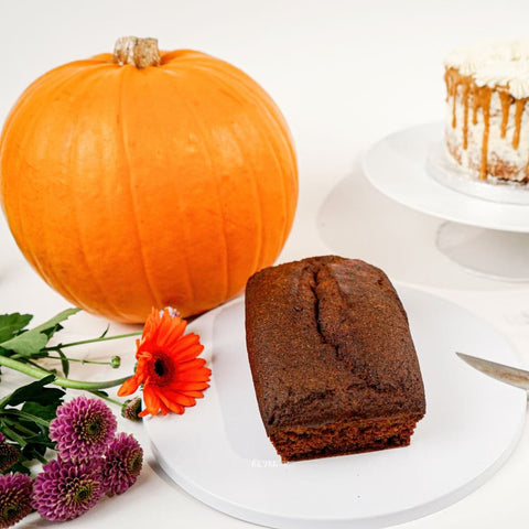 A Pumpkin and Caramel Keto Cake Loaf by No Guilt Bakes, a knife, blooming flowers, and a frosted cake on a stand are arranged on a white surface, complemented by a jar of Caramel Pumpkin Sauce.